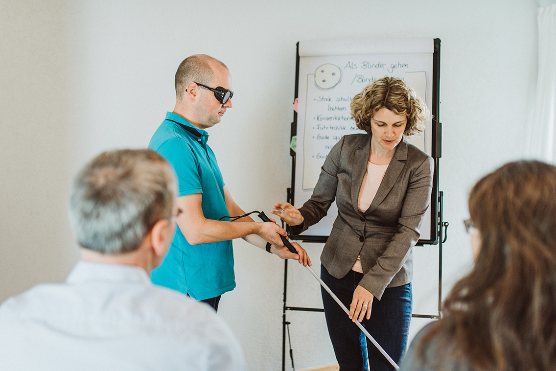 Seminarleiterin Gisela Moser assistiert einem Schulungsteilnehmer mit Simulationsbrille und Langstock